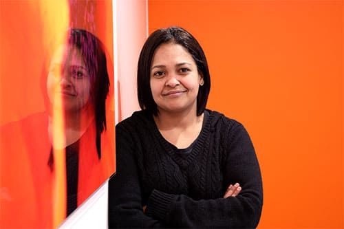 Une femme, les bras croisés, appuyée contre un mur orange, se reflétait dans une œuvre d'art voisine.