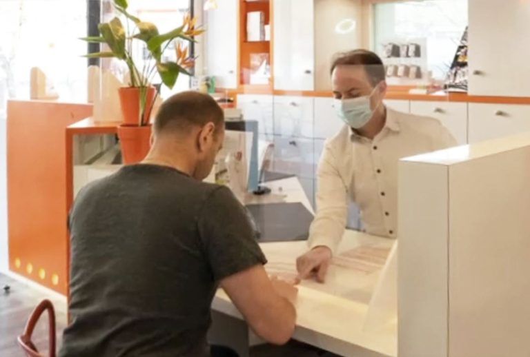 Un homme assis à un bureau parle à une personne portant un masque derrière un comptoir, dans un environnement de bureau.