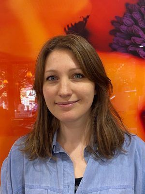 Femme aux cheveux châtain clair, souriante, portant une chemise bleue sur un fond floral orange.