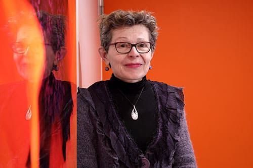 Une femme portant des lunettes se tient près d'un mur orange, vêtue d'un col roulé noir et d'un cardigan violet.
