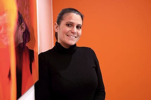 Une femme en col roulé noir sourit, appuyée contre un mur orange orné d'un portrait.