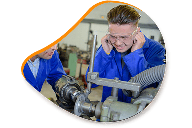 Deux ouvriers dans un atelier ; l’un se protège les oreilles, l’autre travaille sur des machines. Uniformes bleus, lunettes de sécurité.