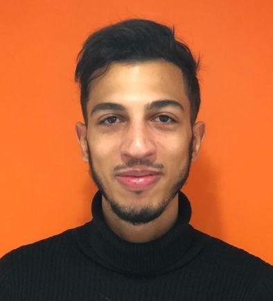 Homme aux cheveux foncés et à la barbe courte, portant un col roulé noir, souriant devant un fond orange.
