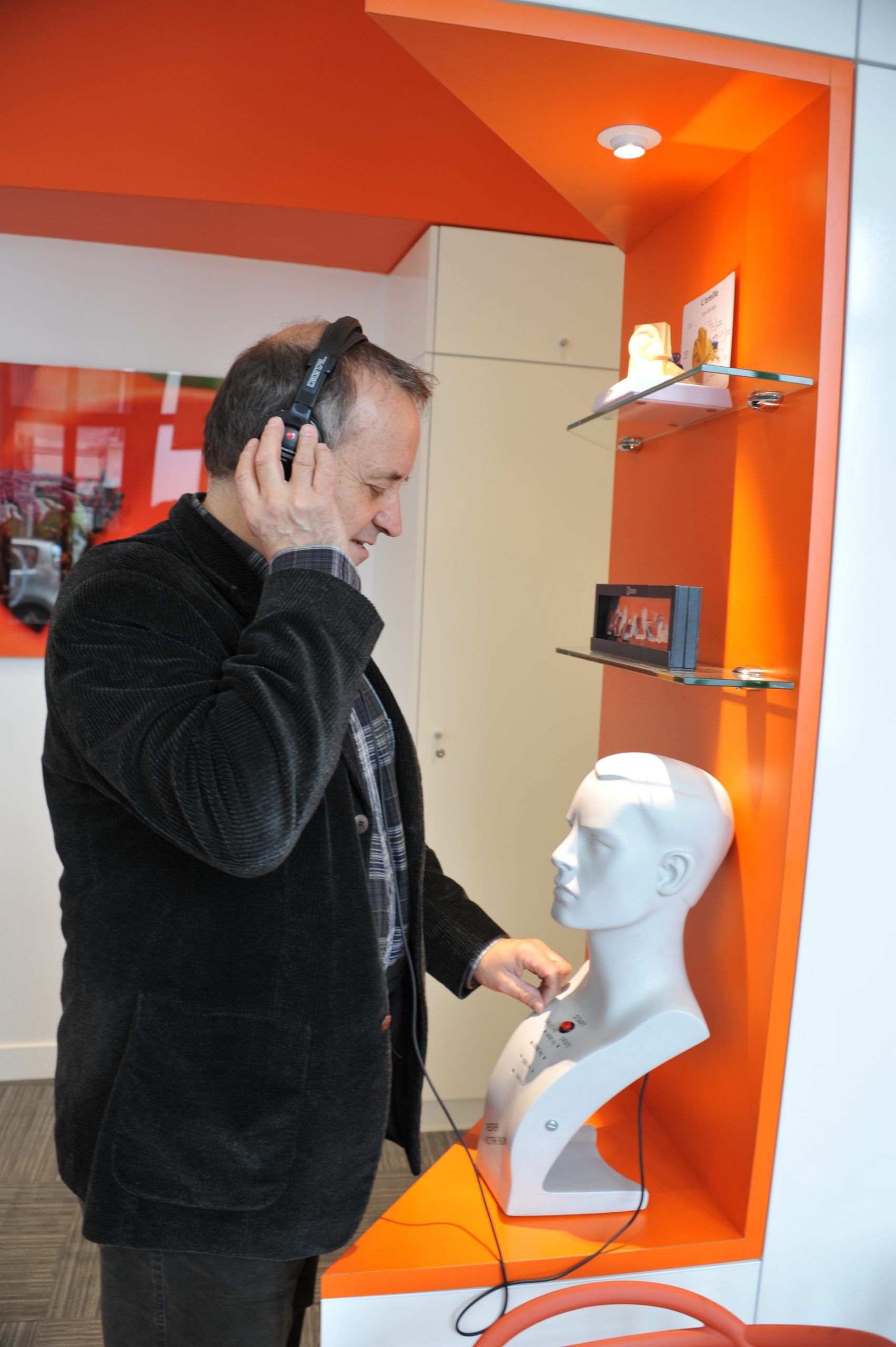 Un homme portant un casque audio écoute un appareil audio relié à une tête de mannequin placée dans une vitrine orange.