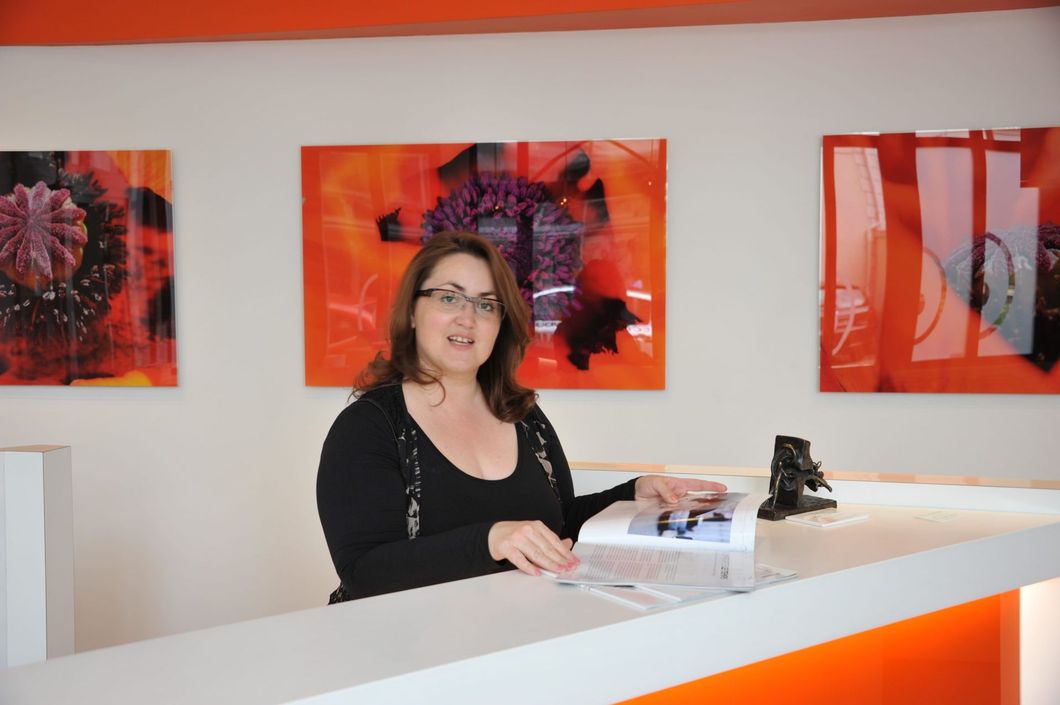 Une femme, assise à un comptoir blanc, désigne une machine. Des œuvres d'art et des touches de couleur orange ornent une pièce lumineuse.