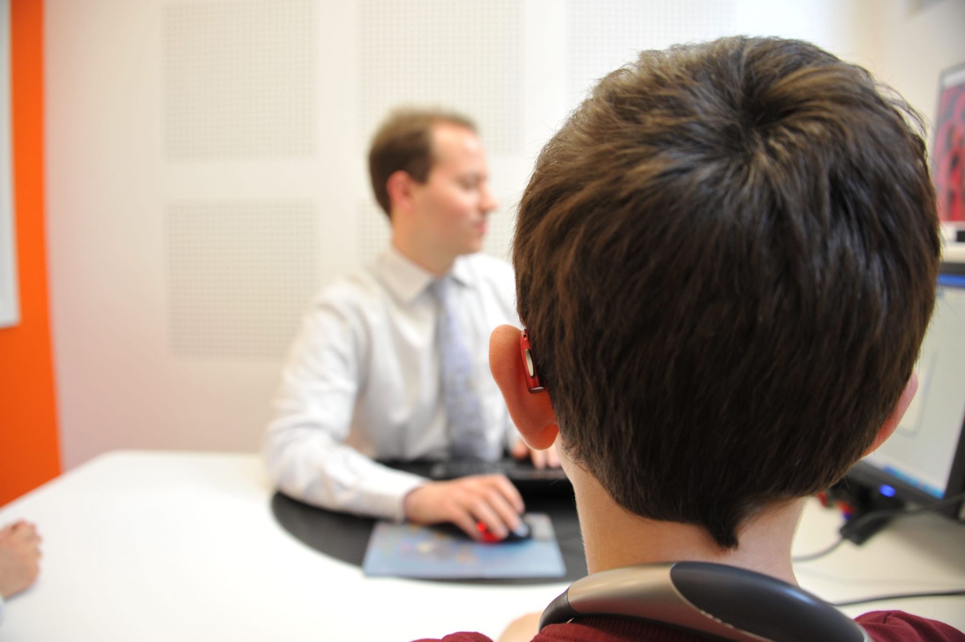 Une personne est assise à un bureau, face à un ordinateur, avec un collègue en arrière-plan. La pièce a des murs orange et blancs.