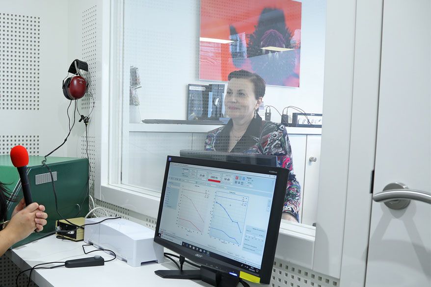 Une femme, assise dans une cabine insonorisée, sourit à travers une vitre. Un écran d'ordinateur affiche des données. Un microphone est visible au premier plan.