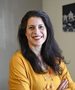 Une femme aux cheveux foncés sourit, les bras croisés, vêtue d'un pull jaune. Photo prise en intérieur.