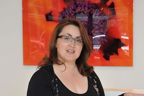 Une femme à lunettes sourit, debout devant une œuvre d'art orange ornée d'éléments violets et noirs.