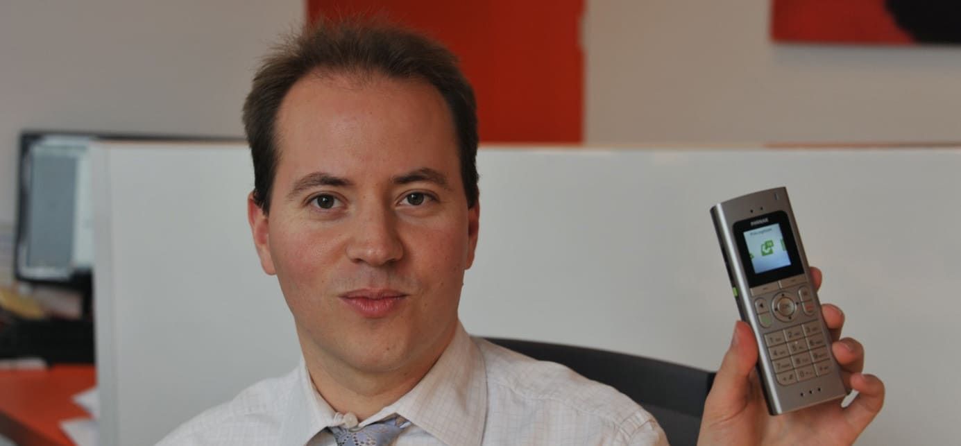 Un homme souriant tient un téléphone portable argenté. Intérieur, fond neutre.