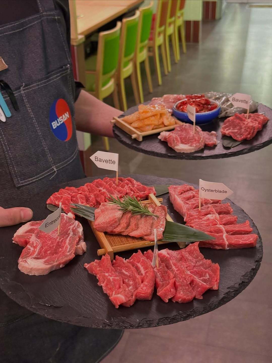 Tray of assorted raw meat cuts at a butcher counter with small labels and garnish upon a person’s apron