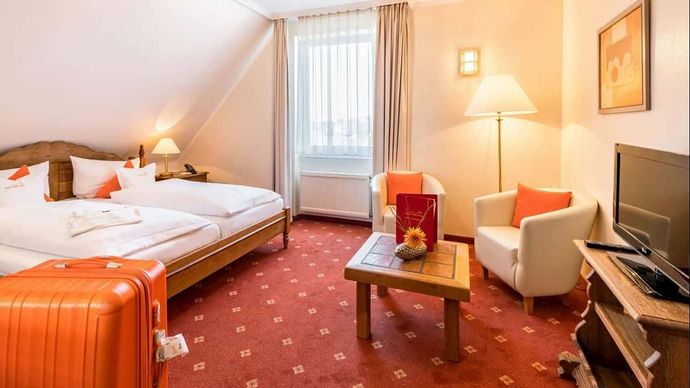 Cozy hotel room with twin beds, orange luggage, armchair, lamp, and TV by a window