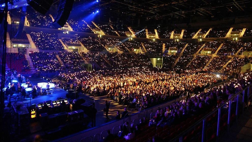 Packed indoor concert arena with a brightly lit stage and a large crowd under blue and gold lights