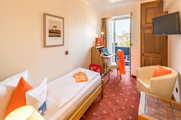 Small hotel room with twin beds, orange accents, desk, and balcony door