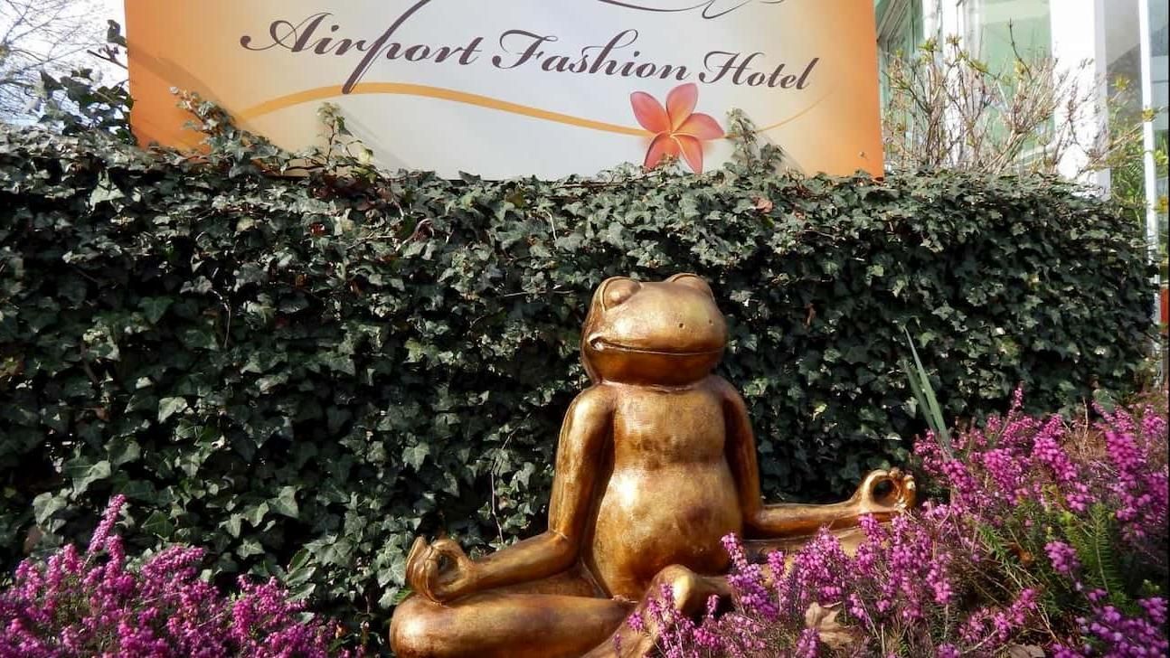 Gold frog statue in a garden with purple flowers and a sign for the Airport Fashion Hotel behind it
