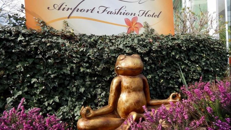 Gold frog statue in a garden with purple flowers and a sign for the Airport Fashion Hotel behind it