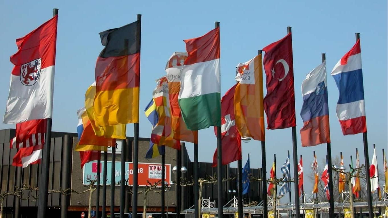 Rows of national flags on poles waving against a blue sky at an outdoor event