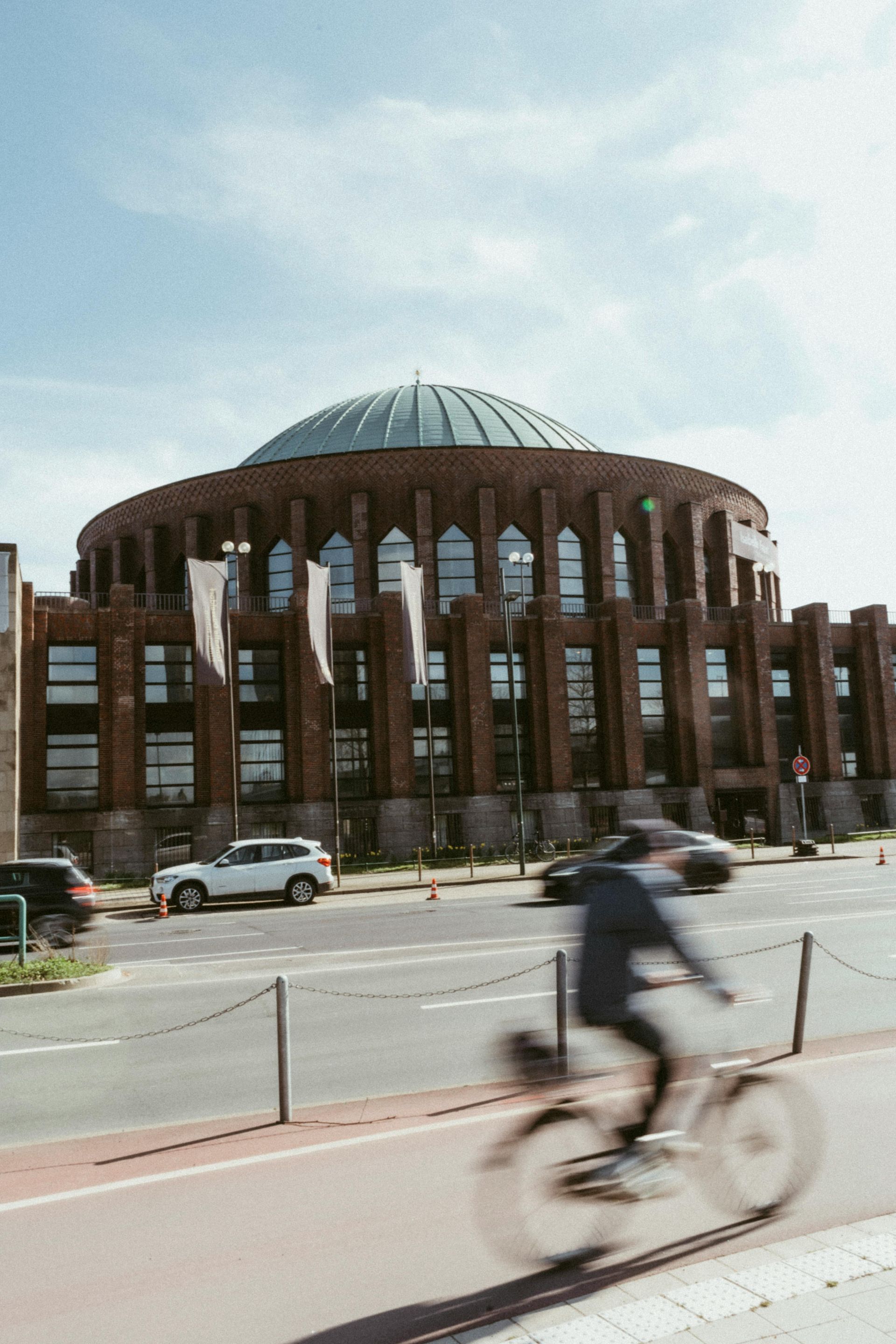 Cyclist blurs past a large circular brick building with a domed roof on a city street.