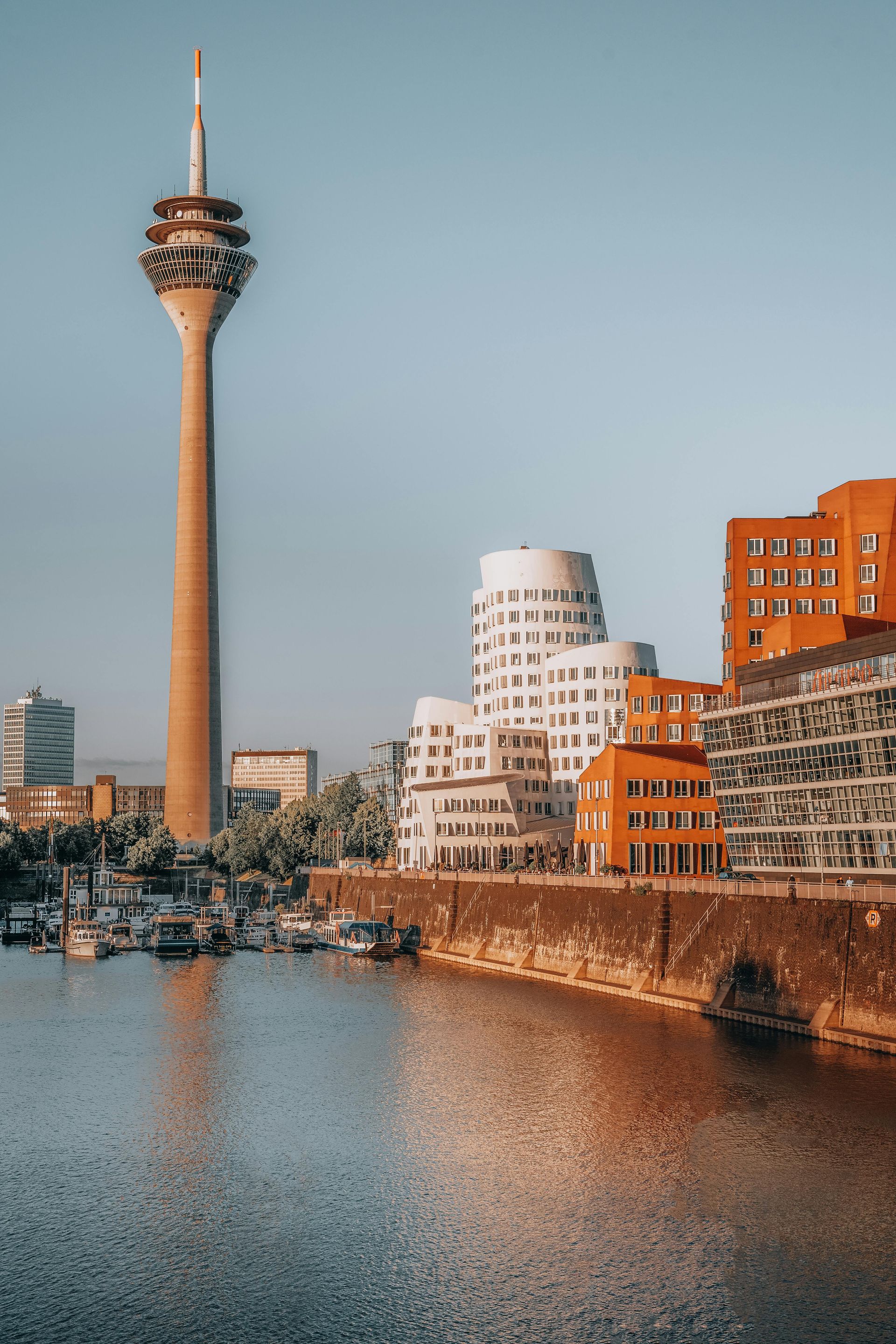 Waterfront city skyline with a tall observation tower, modern buildings, and a calm harbor at sunset