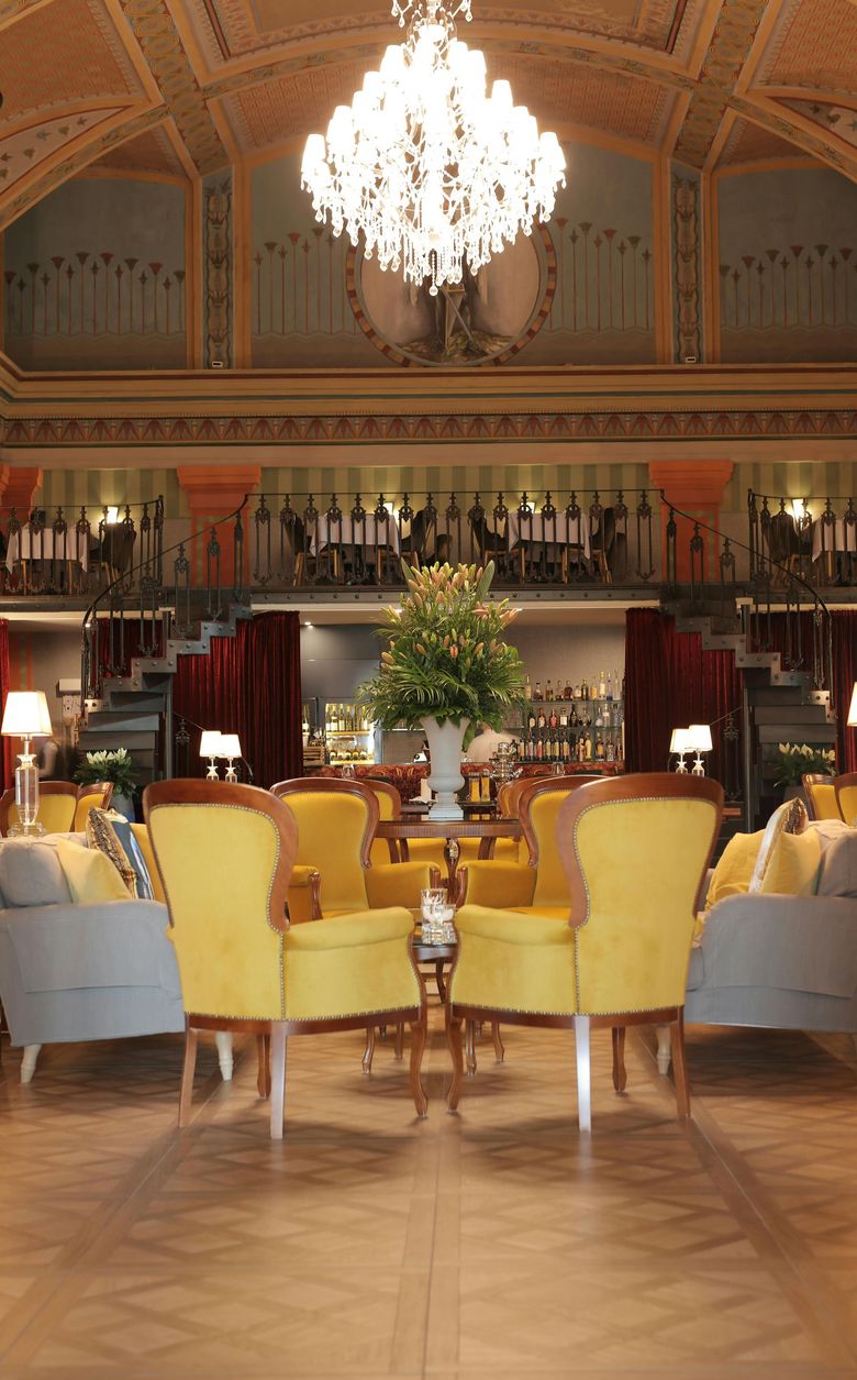 Elegant hotel lounge with yellow armchairs, a chandelier, and a mirrored fireplace at center