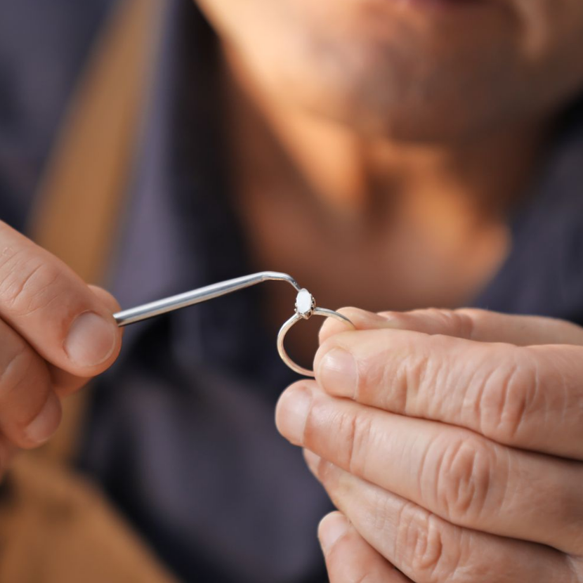 Bijoutier utilisant un outil pour travailler une bague en or ornée d'une petite pierre blanche. Gros plan sur les mains.