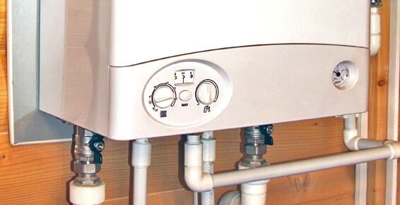 Une chaudière à gaz murale blanche, munie de deux boutons de commande et de tuyaux blancs, installée contre un mur en bois.