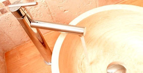 Dans une salle de bains carrelée, un robinet chromé distribue l'eau dans un lavabo rond en pierre beige.