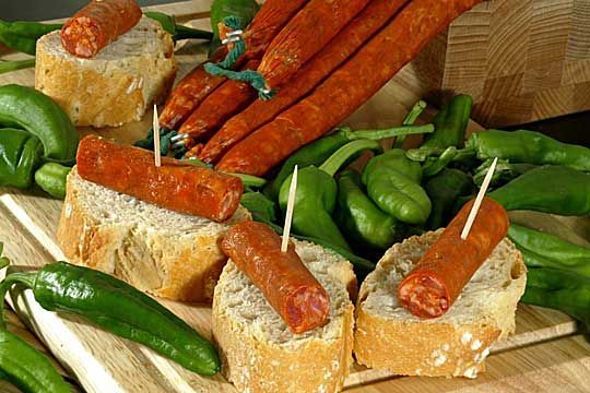 Una tabla de cortar de madera cubierta con pan, pimientos y salchichas.