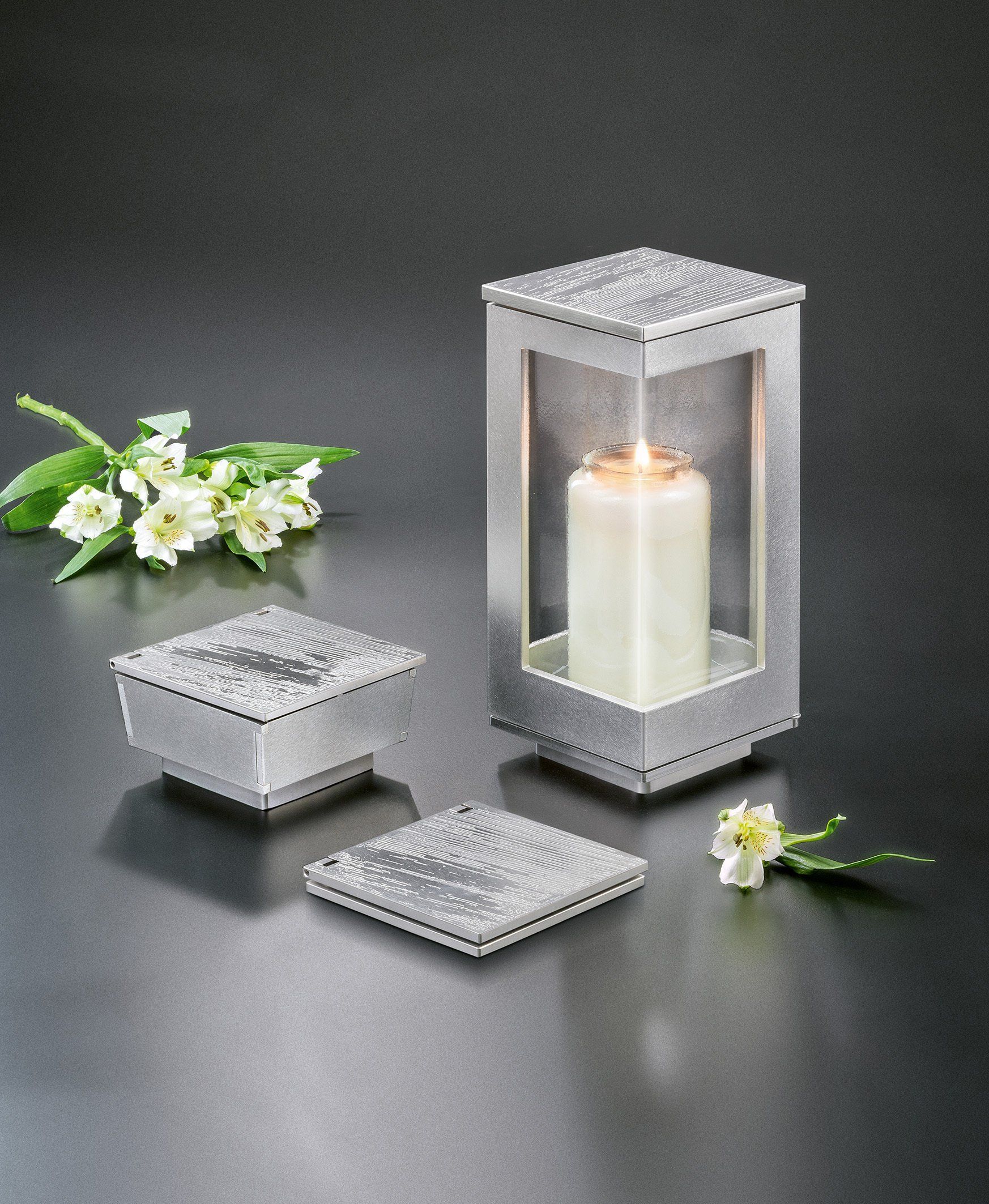 Silberne Kerzenlaterne mit brennender Kerze, zwei kleinen silbernen Schachteln und weißen Blumen auf dunklem Hintergrund.