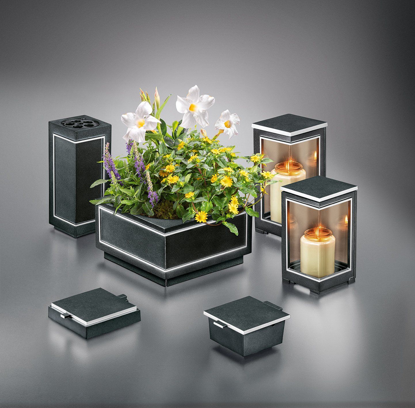 Ein dunkles Blumenarrangement aus Granit und Kerzenhalter mit Blumen und brennenden Kerzen vor einem grauen Hintergrund.