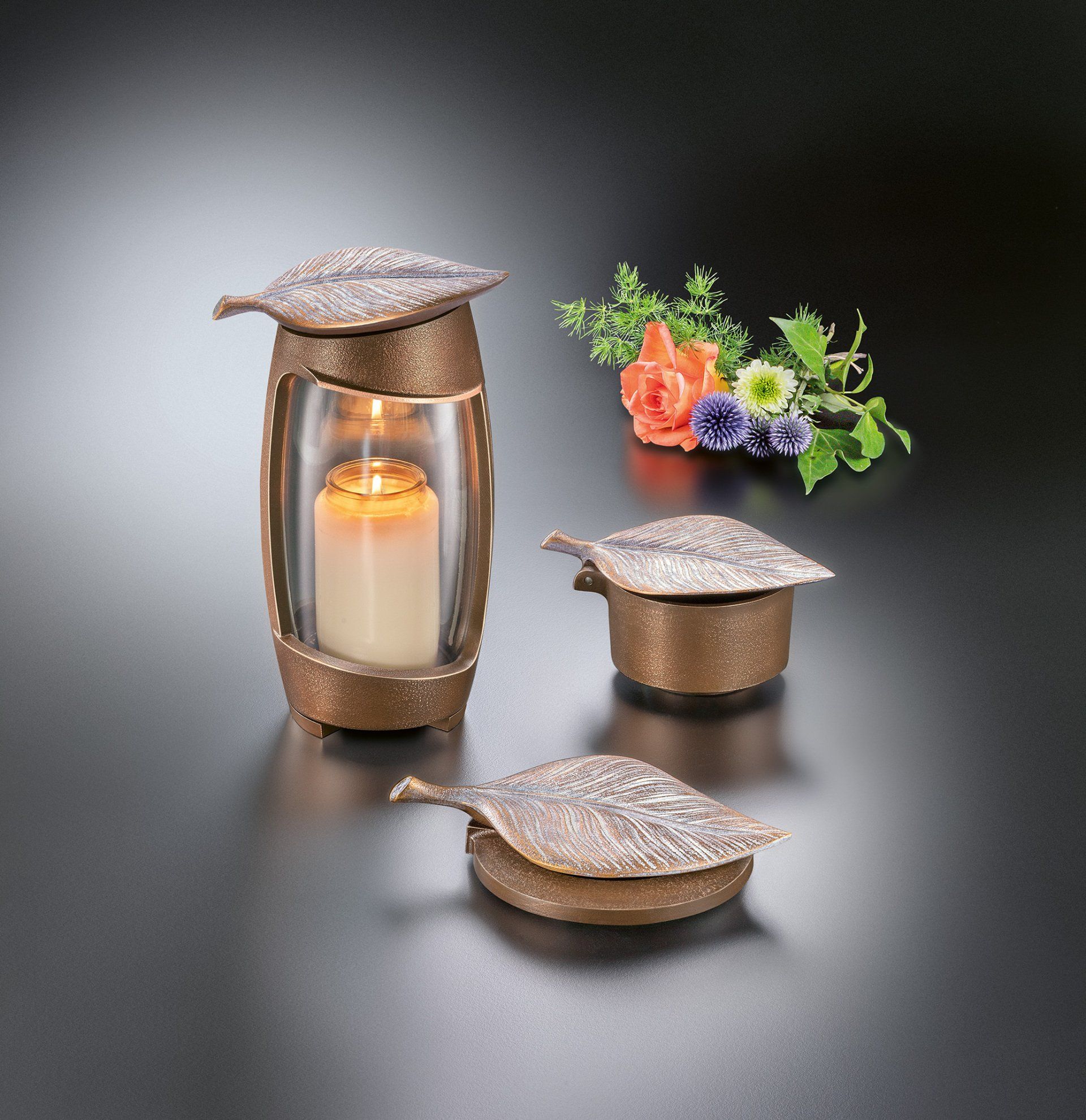 Kerzenhalter in Blattform aus Bronze, kleine Schachtel und Urne mit brennender Kerze darin, Blumen auf schwarzer Oberfläche.