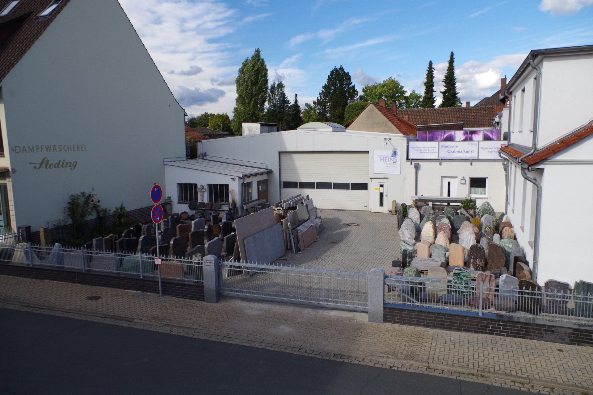 Ein Steingeschäft mit verschiedenen Grabsteinen und Gebäuden, gepflastertem Grundstück und einem bewölkten blauen Himmel.