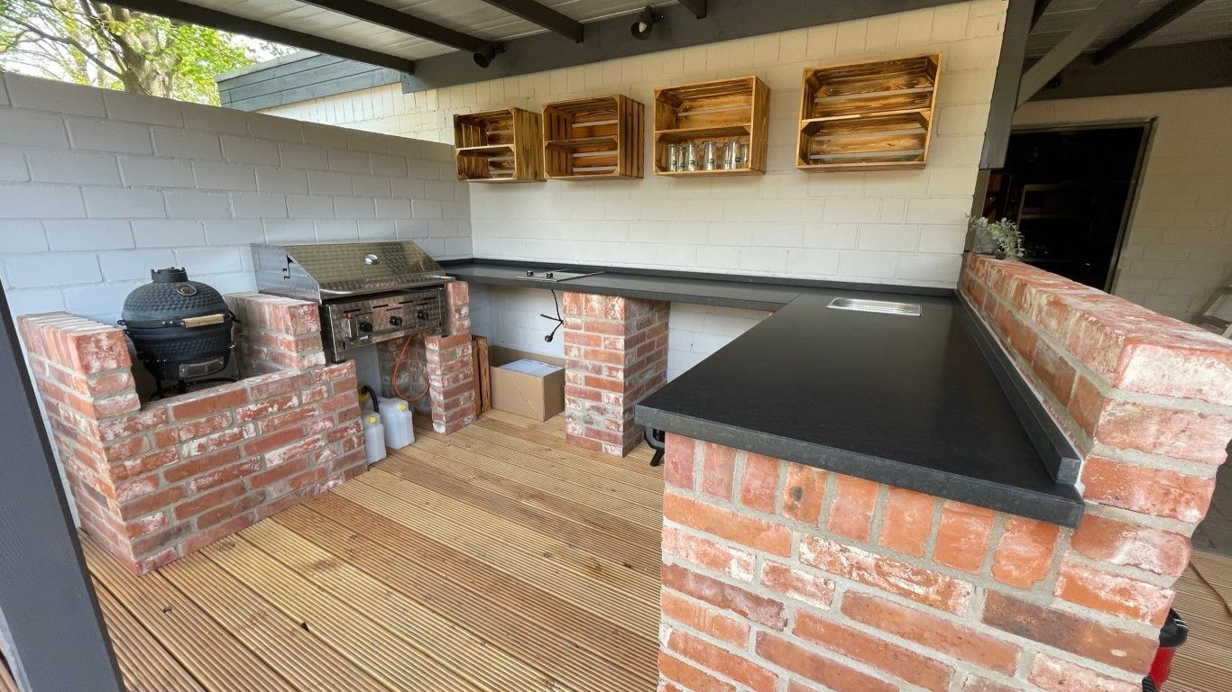 Außenküche mit Ziegel- und Holzelementen, schwarzer Arbeitsplatte, Grill und Regalen.