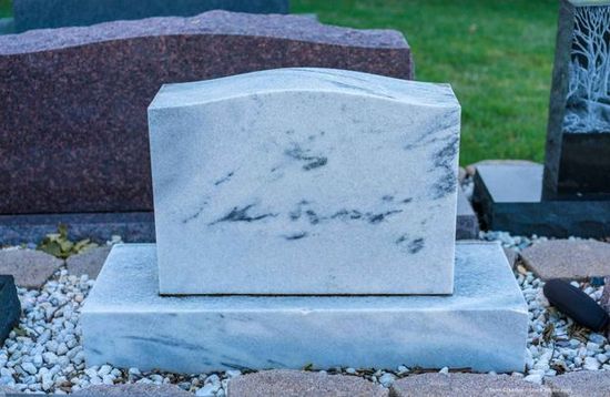 Grabstein aus weißem Marmor mit dunkelgrauer Maserung auf einem rechteckigen Sockel auf einem Friedhof.