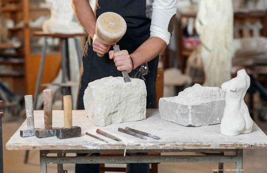 Bildhauer meißelt mit Hammer und Meißel an einer Werkbank Stein, umgeben von Werkzeugen und gemeißelten Figuren.