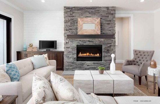 Wohnzimmer mit Steinkamin, hellbraunem Sofa, grauem Sessel und Couchtisch aus Holz.