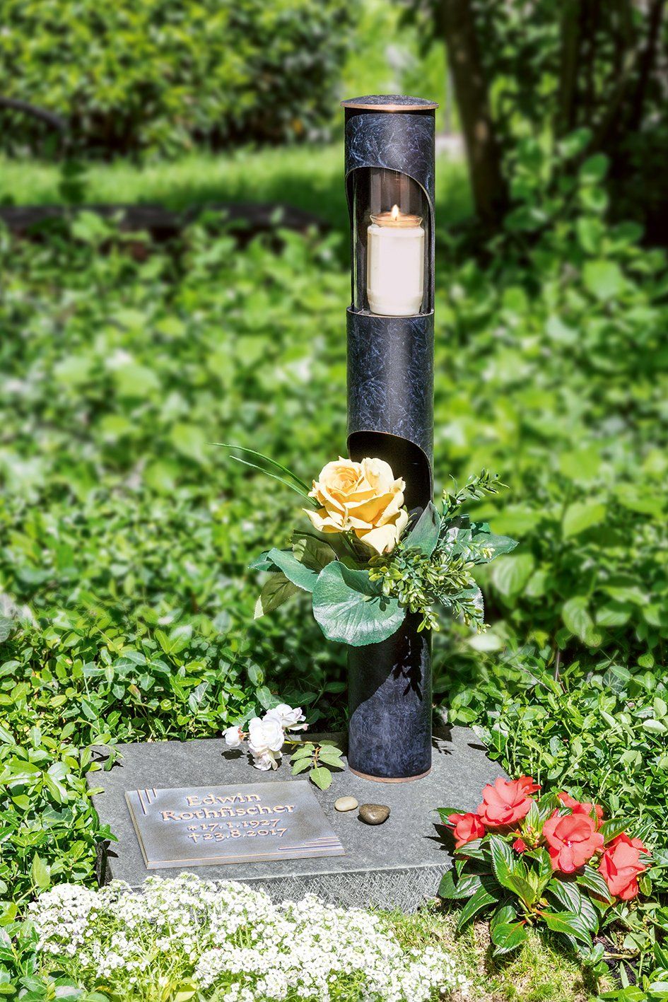 Grabstein mit Kerzenhalter, gelber Rose und roten Blumen. In einer grünen Gartenumgebung.