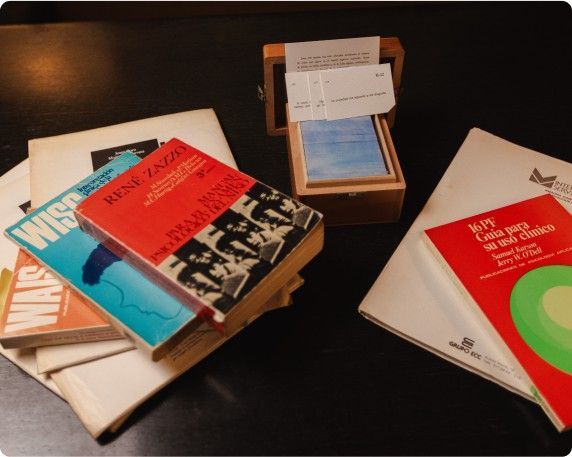 Pilas de libros y una pequeña caja expositora de madera sobre una mesa oscura, con portadas coloridas y tarjetas a la vista.