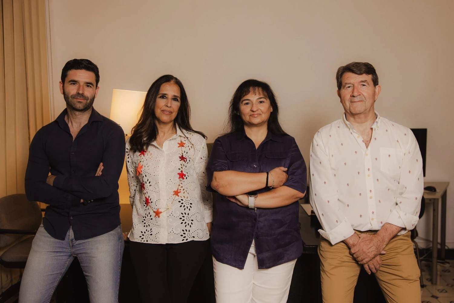 Cuatro personas de pie en un interior, posando con los brazos cruzados frente a una pared lisa.