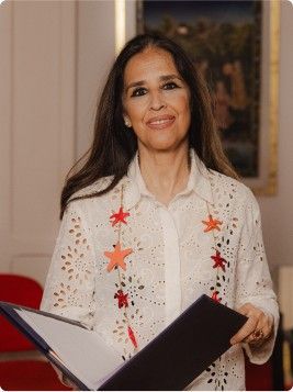 Mujer sonriendo y sosteniendo una carpeta negra abierta, con una blusa blanca bordada y adornos de estrellas rojas.