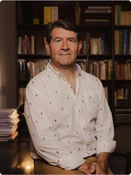 Hombre sentado frente a unas estanterías, con una camisa blanca estampada, en una habitación con una iluminación cálida.