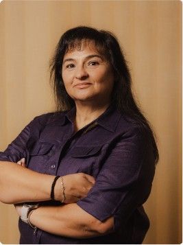 Persona con una camisa morada y los brazos cruzados, de pie frente a un fondo beige.