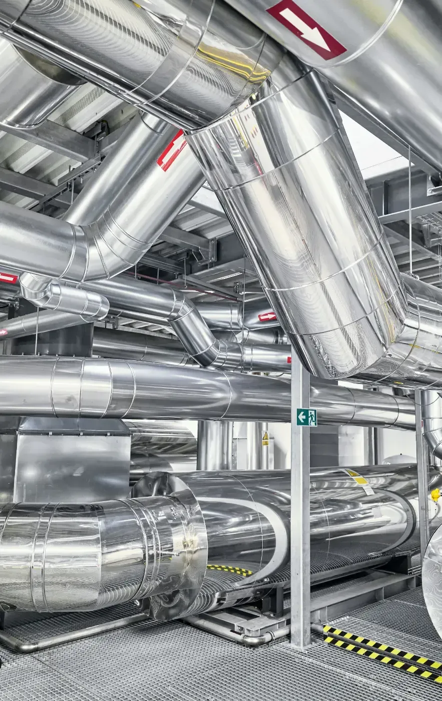 Glänzende silberne Industrierohre und -kanäle in einem Maschinenraum, mit Deckenisolierung und Metallträgern.