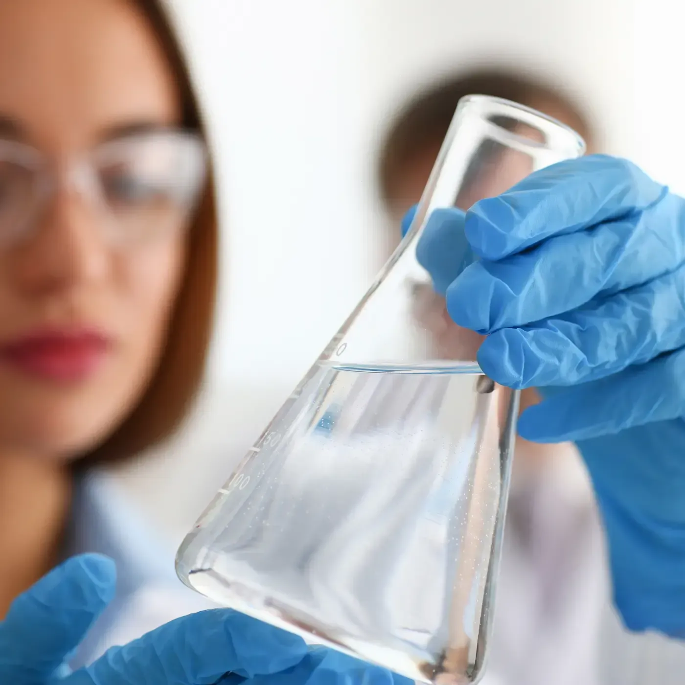 Behandschuhte Hände halten einen durchsichtigen Laborkolben, während im Hintergrund ein verschwommener Wissenschaftler zusieht.