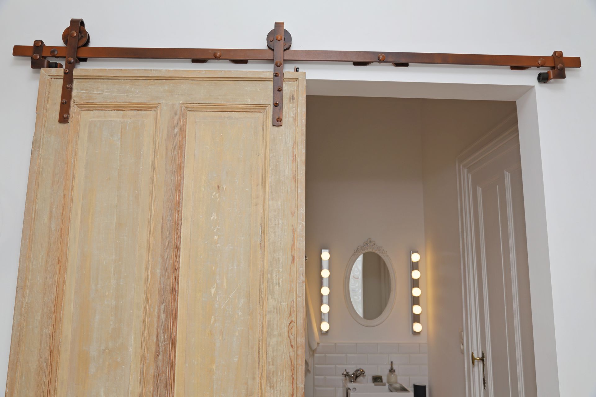 Une porte coulissante en bois clair, avec des ferrures en bronze, s'ouvre sur une salle de bains avec miroir et éclairage.