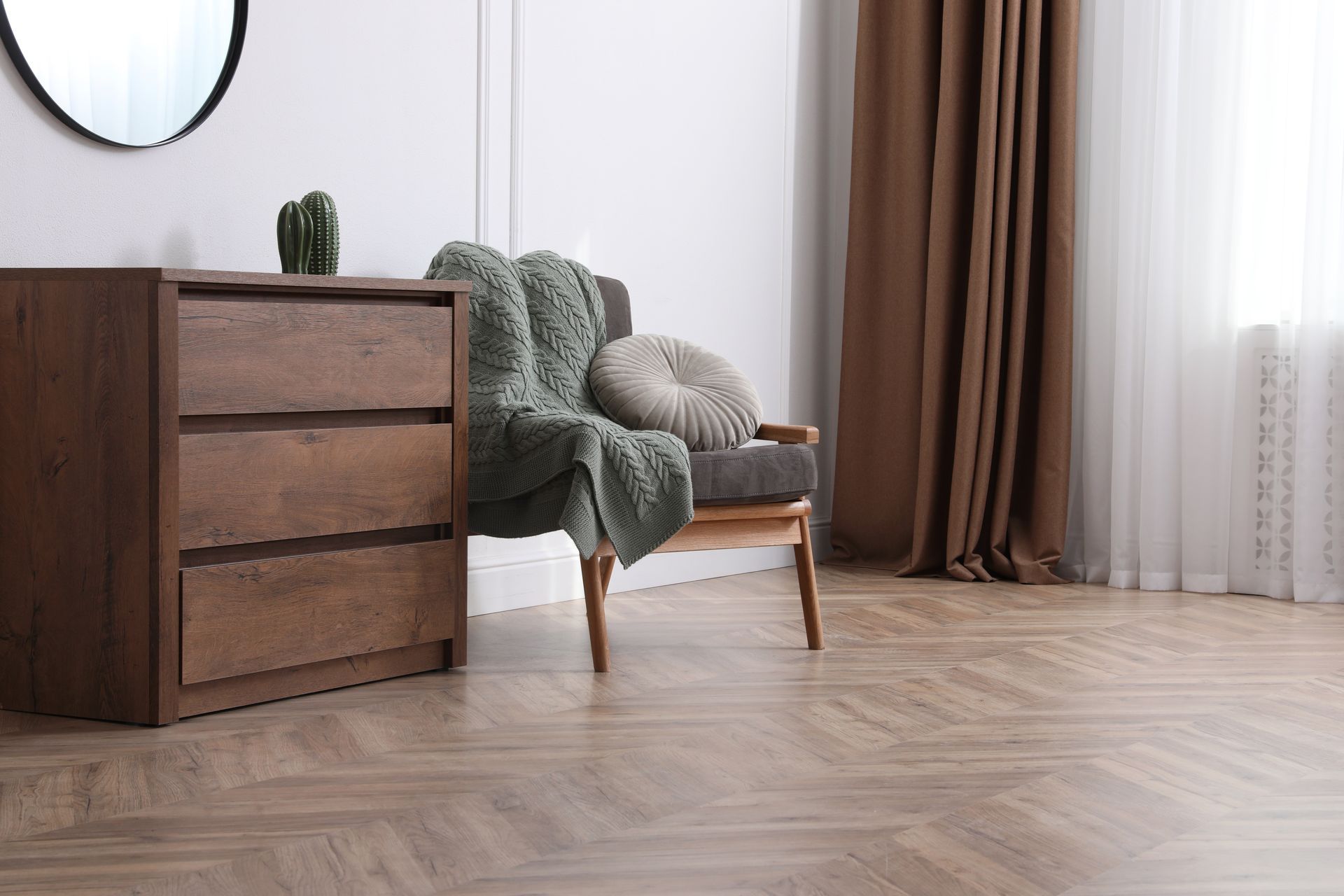 Une commode en bois, un fauteuil gris avec une couverture et des coussins, des rideaux marron et un miroir rond dans une pièce.