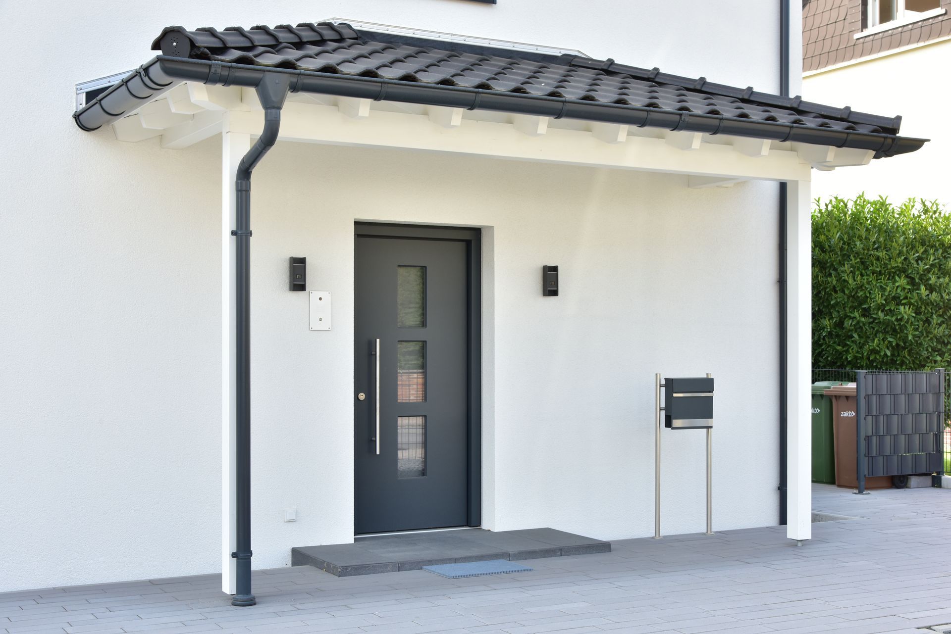 Entrée de maison blanche avec porte noire, auvent et boîte aux lettres. Toit gris, boiseries blanches.