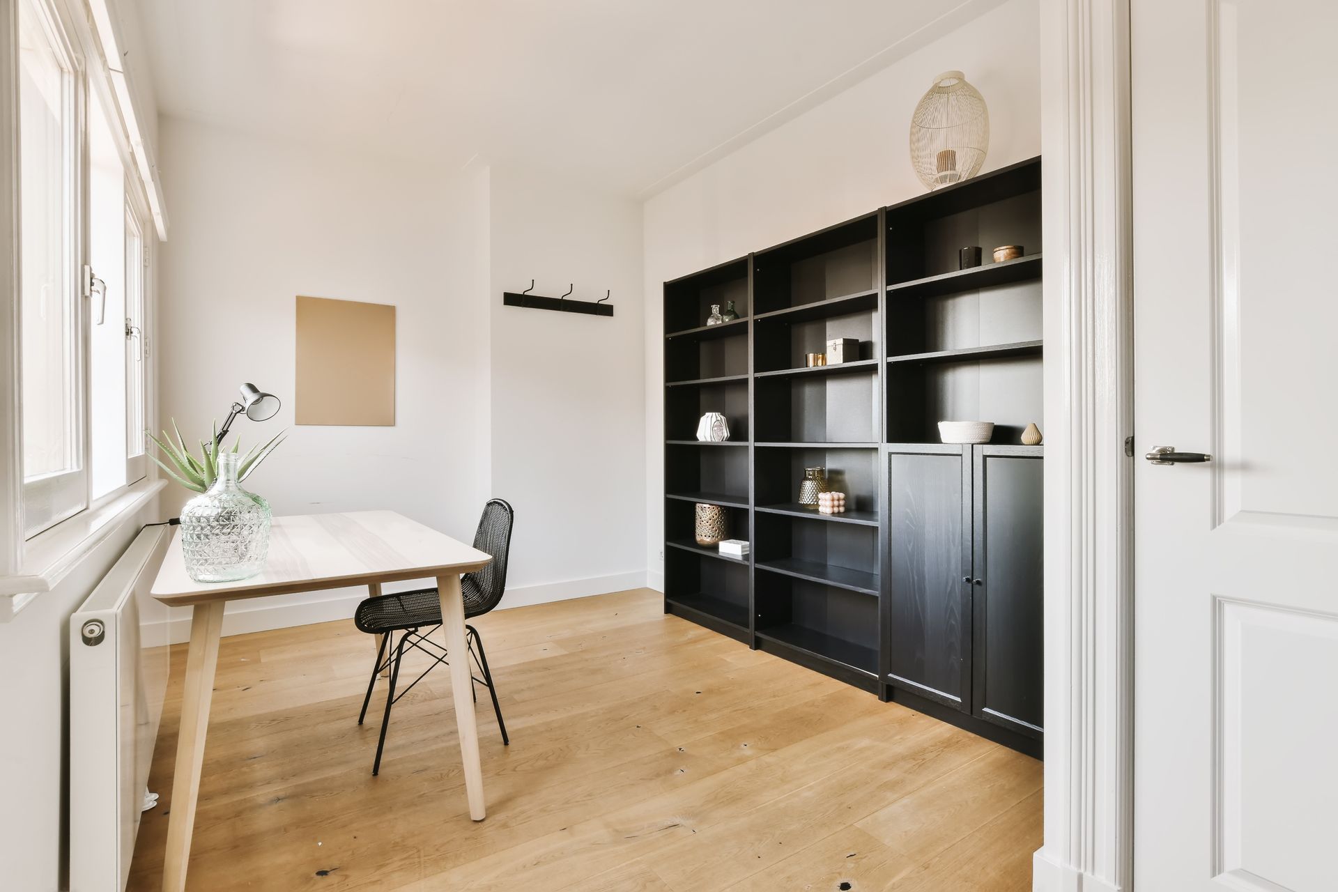 Un bureau à domicile avec un bureau, une chaise, une bibliothèque noire intégrée, des murs et une porte blancs et un parquet clair.