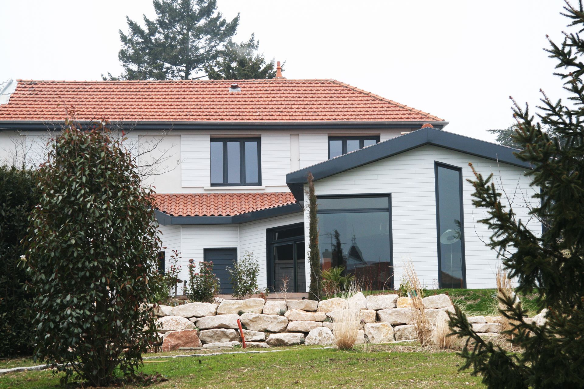 Vue extérieure d'une maison blanche moderne aux encadrements de fenêtres et volets gris foncé. Pelouse et jardin verdoyants.