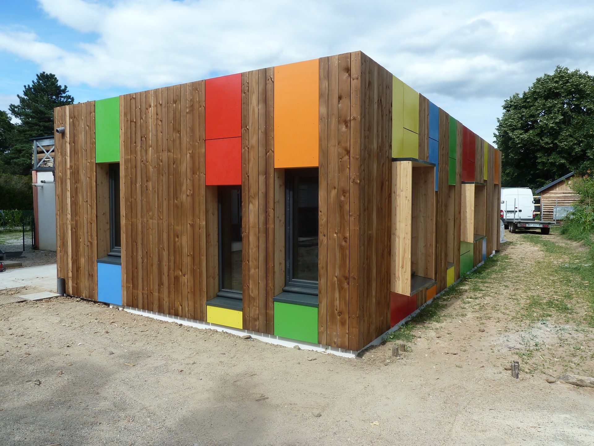 Bâtiment en bois avec des panneaux colorés autour des fenêtres, sur un terrain gravillonné sous un ciel nuageux.